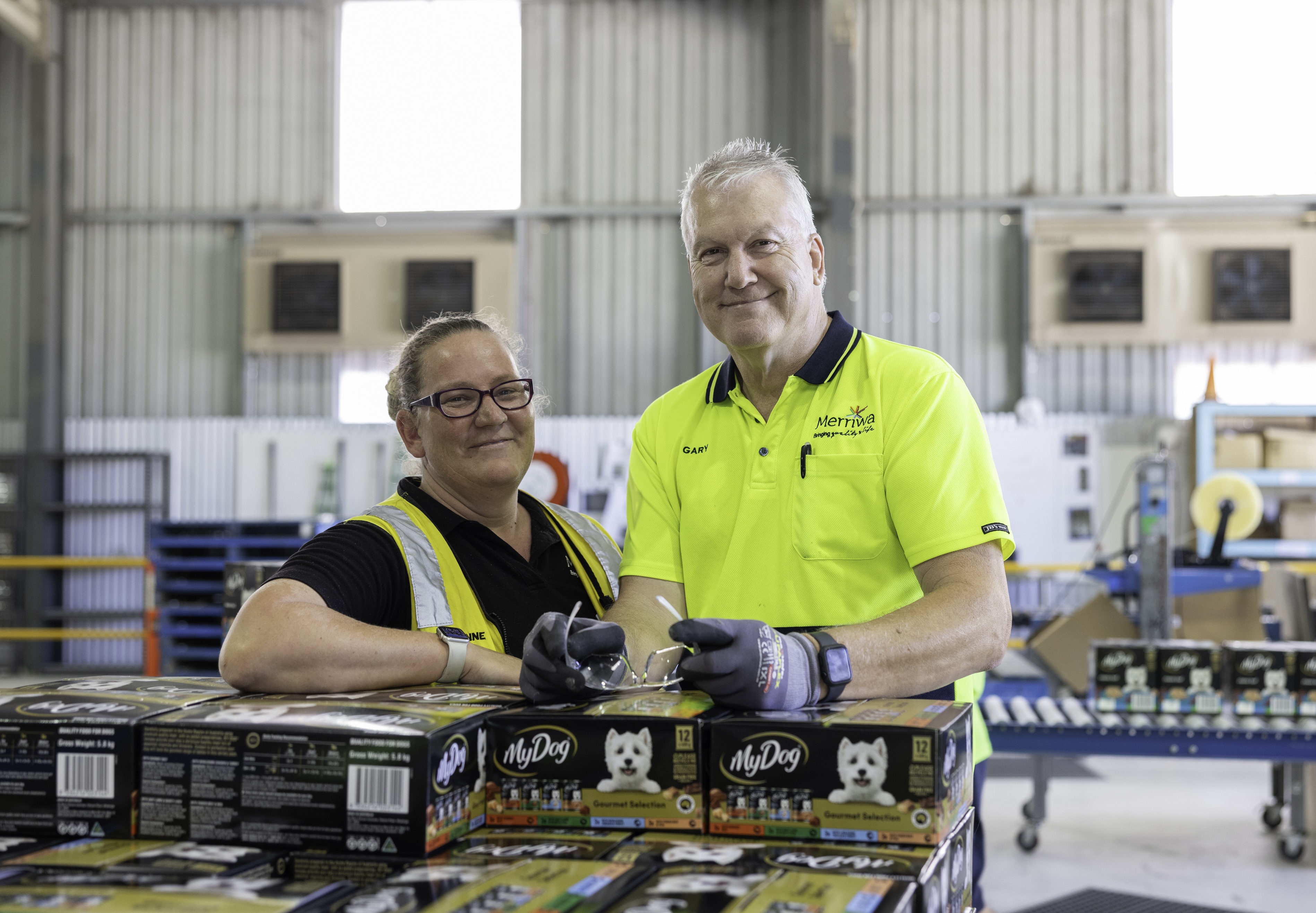 Two warehouse workers in hi-vis uniforms smile while standing beside stacks of boxed pet-food products.
